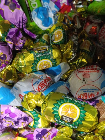 The image is a close-up, overhead shot of a pile of individually wrapped candies. The wrappers are a variety of colors, including gold, purple, green, white, and blue. Some wrappers feature images of fruit slices, particularly lemons, and some have Cyrillic text. One prominent candy wrapper is gold with a green border and a yellow lemon slice illustration, with the text "BAYAN SULU Лимонный" visible. Another wrapper is white with a red circular design and a cartoon cow illustration, also with Cyrillic text. The overall impression is a colorful and abundant collection of sweets. There are no people, actions, or interactions depicted. The environment is not discernable beyond the candies themselves. There is no information in the image that allows for the determination of its location in the city of Martuni, Armenia.