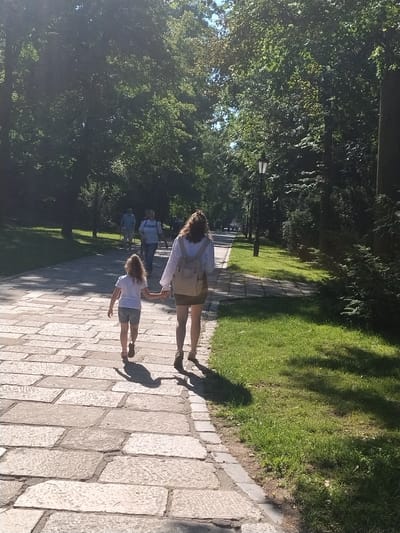 The image depicts a paved walkway in a park, lined with trees and grass. In the foreground, a woman and a young girl are walking away from the camera, holding hands. The woman is wearing a light-colored top, a dark skirt, and a backpack, while the girl is in a white t-shirt and shorts. Further down the path, other individuals are visible walking in the distance. The scene appears to be a sunny day, with dappled sunlight filtering through the trees. The location is identified as the Velkopopovická street in Písek, Czechia.