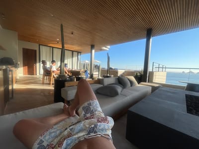 A first-person perspective photograph captures a partially-clad individual reclining on a cushioned lounge area in an open-air, upscale setting. The subject's torso and legs, clad in patterned swim trunks, are prominently featured. The lounge area includes a substantial grey couch with multiple pillows and a dark-colored fire pit table. In the background, two cactus plants in pots stand between the lounge area and a dining setup where three individuals are seated. Beyond the immediate indoor/outdoor space, the view opens up to a railing and steps leading down to a coastal scene with ocean and rock formations visible. The architecture features a slatted wood ceiling. The location is inferred to be Cabo San Lucas, Mexico, based on the landscape and architectural style.