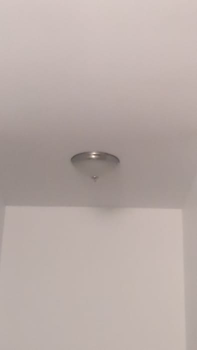 The image shows a ceiling light fixture in a room in Roxton Pond, Canada. The fixture has a white frosted glass shade and a silver metal frame. It is mounted in the center of the ceiling. There are no people in the room. The image captures the ceiling and part of two walls. The walls are painted white. The ceiling is white and has a slight texture. There is no furniture or other objects in the room.