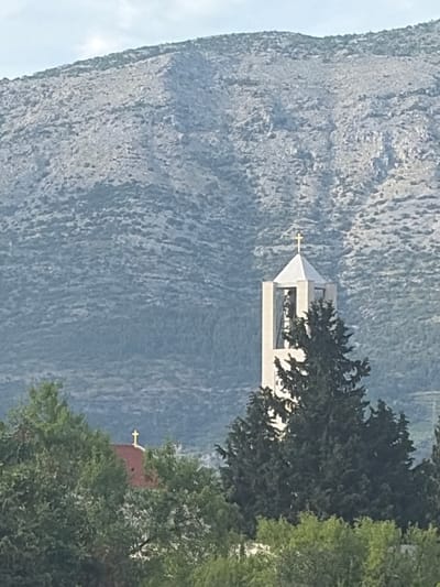 The image displays a daytime scene in Grad Korčula, Croatia, dominated by natural landscape and religious architecture. In the foreground, dense green deciduous foliage occupies the lower portion of the frame. Directly behind this, a light-colored, square-based bell tower with a white pyramidal roof, crowned by a golden Latin cross, stands prominently. The tower's upper section features an open belfry, partially fronted by a large, dark green coniferous tree. To the left and slightly below the bell tower, a section of a red-tiled roof, also topped with a smaller golden Latin cross, is visible amidst the surrounding green canopy. The entire background is occupied by a large, rugged mountain characterized by a grey, rocky surface sparsely covered with low, scrubby vegetation. The sky above the mountain is light blue and appears hazy. No individuals, discernible actions, or direct interactions are depicted in the static scene.