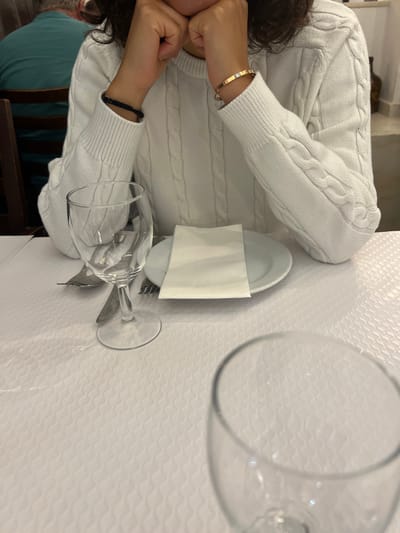 The image depicts an indoor dining scene in Coruche, Portugal. In the foreground, a person wearing a white cable-knit sweater is seated at a table covered by a white textured tablecloth. Their hands are clasped near their lower face. A black cord bracelet is on their left wrist, and a gold-toned bangle with a single square stone is on their right wrist. The table setting includes a white ceramic plate with a folded white paper napkin, a metal fork, and a knife. A clear stemmed wine glass is positioned left of the plate. The rim of a second clear glass is partially visible in the immediate foreground. In the blurred background, a second individual wearing a teal shirt is visible, along with wooden dining chairs and light-colored walls, indicative of a restaurant setting.