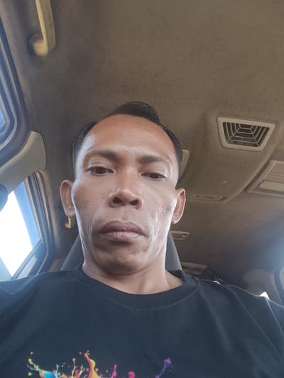A close-up selfie of a man inside a vehicle. The man is looking directly at the camera, with his face filling most of the frame. He has dark hair, brown eyes, and is wearing a black t-shirt with a colorful, abstract design on the front. He is also wearing a gold hoop earring in his left ear. The interior of the vehicle is visible in the background, including the headliner and an air vent. The lighting suggests it is daytime. The exact location in Tiang Tara, Indonesia cannot be determined from the image alone.