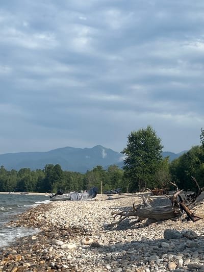 A rocky shore of a body of water is depicted with the water lapping at the stones. In the foreground, a large piece of driftwood rests on the shore. Further back, several inflatable boats with engines are visible, some covered with tarps. A dense forest line runs along the horizon, with mountains visible beyond the trees. The sky is overcast with clouds. This scene is located in the city of Tolbazikha, Russia.