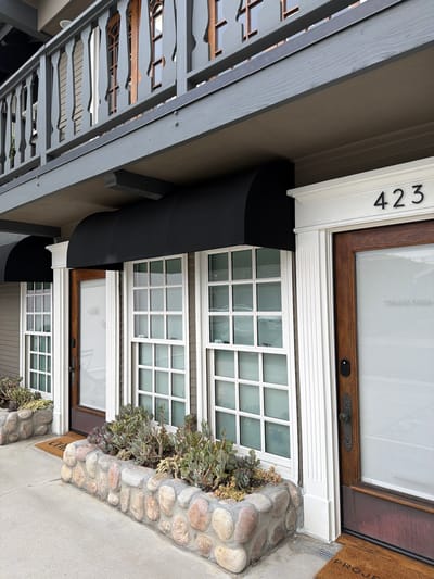 The image captures the exterior of a building located at 423 in Newport Beach, California. The architecture features a front door and adjacent windows with white grilles. A black awning extends over the windows. Stone landscaping is present near the building's entrance. A grey, decorative porch roof or balcony overhangs the entrance area. The photograph appears to be taken during daylight hours.
