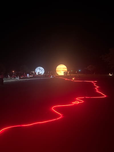The image depicts an outdoor nighttime scene in Baan Pang Ka, Thailand, featuring two large, illuminated inflatable spheres resembling planets or moons. The sphere on the left is textured to look like the moon, while the one on the right is yellow with stripes, possibly representing Jupiter. Numerous people are gathered around these illuminated objects, some standing and others sitting. In the foreground, a bright red neon light rope snakes across the ground, creating a winding path. The ground appears to be a dark, somewhat sandy or gravelly surface. The background is dark, indicating nighttime, with some trees visible on the right side. The overall atmosphere suggests an art installation or event taking place outdoors.