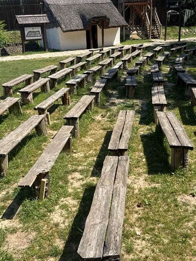 An outdoor amphitheater with rows of weathered wooden benches is set on a grassy area. A rustic building with a thatched roof and white walls is visible in the background, along with a wooden walkway and stairs. Signage indicates the location as "IHDJA" and "#kulturno_selo". The scene suggests a historical or cultural heritage site within Indjija Municipality, Serbia, likely prepared for performances or gatherings. No people are present in the image.