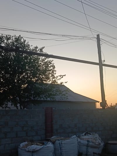 The image shows a dusk scene with a clear sky, transitioning from pale blue to a soft orange hue near the horizon. Several power lines are visible, crisscrossing the upper portion of the frame. A single, weathered wooden utility pole stands on the right side, with electrical wires attached. In the middle ground, a low-slung building with a gray, corrugated roof is partially obscured by a silhouetted tree with dense foliage on the left. In the foreground, a low wall constructed from light gray cinder blocks runs across the bottom of the image. To the right of a red brick column integrated into the wall, three large, white, woven bags are piled up, appearing to contain loose material, possibly sand or construction debris. The overall impression is of a rural or semi-urban setting in Altinkul, Uzbekistan, during twilight.