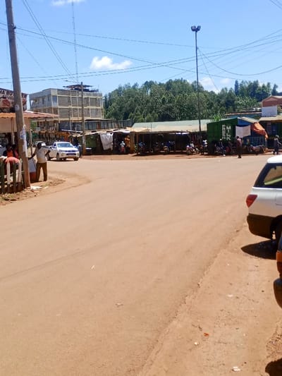 The image depicts a daytime scene in a commercial area of Kahuro, Kenya, under a clear blue sky with sparse clouds. The foreground consists of an unpaved, reddish-brown road surface, with the rear of a white sedan partially visible on the right. In the midground, a white pickup truck traverses left to right. To the left, a man in a light shirt and dark trousers stands near a wooden fence and a utility pole with overhead power lines. A multi-story concrete building with exposed rebar and multiple windows is visible behind him. On the right, a line of rudimentary market stalls constructed from wood, tarpaulins, and corrugated metal sheets, including a green shipping container-like structure labeled "FRESH MILK," borders the road. Numerous motorcycles are parked at these stalls, with several individuals standing or sitting among them. A tall street light with multiple floodlights stands above the stalls. Lush green foliage and trees are visible on a hillside in the background. Several other people are scattered throughout the market area, engaged in various, indistinct activities.