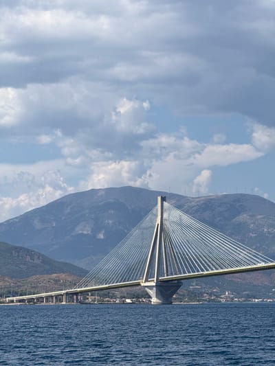 This media file depicts a cable-stayed bridge spanning a body of water, likely a strait or bay, with a mountainous coastline on one side and the open sea on the other. The bridge is the dominant object, with its tall, slender pylons and numerous cables creating a striking geometric pattern against the sky and mountains. The sea is a deep blue with a rippled texture, suggesting it is somewhat rough or windy.
The scene is set during the day, under a partly cloudy sky with patches of blue visible between the white and gray clouds. The mountains in the background are rugged and covered in sparse vegetation, with a small settlement visible along the coastline. Several tall, thin structures, possibly electricity pylons or wind turbines, are lined up along the crest of the mountain.
The perspective is from a viewpoint slightly above the water, looking towards the bridge and the coastline. The image's orientation is rotated, showing the scene sideways. No people are visible in the frame, and no text is present. The overall impression is one of impressive engineering set against a dramatic natural landscape. The location context indicates this is likely the Rio-Antirrio Bridge in the Municipal Unit of Rio, Greece, which connects the Peloponnese peninsula with mainland Greece over the Gulf of Corinth.