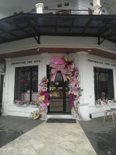 The image displays the exterior of a commercial establishment, likely a bakery or cafe, situated in the city of Bojongherang, Indonesia. The building features a white facade with dark trim, an angled front, and a dark, transparent canopy extending over the entrance. A white decorative pillar or structure is visible on the roofline. The central entrance consists of double glass doors framed in black, adorned with extensive garlands of multicolored artificial flowers, including shades of pink, purple, yellow, and red, which cascade from the top and sides, with some flowers resting on the pavement. A large pink and white striped bow is prominently displayed above the doors. A pink neon sign reading 'Katumbah Sweet Home' is located above the bow. Large display windows flank the entrance; the left window is labeled 'CONNECTING THE LOVE,' and the right window is labeled 'PERSONALIZE YOUR CAKE.' Both windows feature flower boxes on their ledges. Inside the establishment, through the glass, baked goods are visible on display shelves. On the tiled grey pavement to the right of the entrance, two modern white chairs with grey cushions are positioned. The pavement exhibits damp patches, particularly on the left side. No individuals are present in the scene.