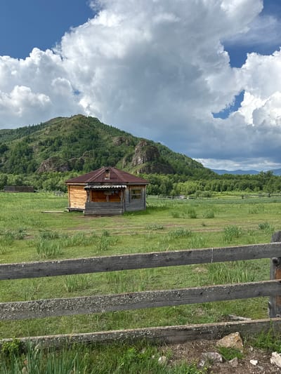 An outdoor, eye-level, medium shot captures a rustic wooden cabin nestled in a grassy field, with a lush, green mountainous backdrop. The sky is a dramatic mix of blue patches and billowing white clouds. In the foreground, a weathered wooden fence, composed of horizontal planks, runs across the bottom of the frame, partially obscuring the view of the field.
The cabin, located in the village of Kazanovka, Russia, is constructed from light-colored wooden logs. It features a prominent, sloping roof made of corrugated metal, with a small chimney. A covered porch extends from the front of the cabin, with a few items of clothing hanging on a line. The cabin's windows are visible, with one appearing to have a glass pane. The surrounding field is covered in tall, green grass and some scattered vegetation.
Behind the cabin, a wooden fence, similar in style to the one in the foreground, encircles a portion of the field. Beyond the fence, the landscape rises into a verdant, tree-covered mountain. The mountain slopes are densely packed with various shades of green foliage, with rocky outcrops visible. The overall scene evokes a sense of rural tranquility and natural beauty.