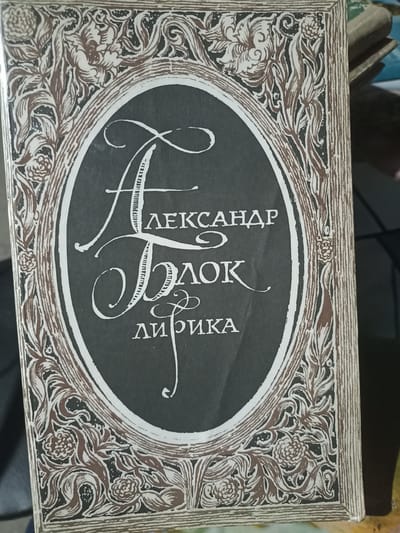 The image shows the cover of a book with a decorative border and an oval frame containing text. The text appears to be in Russian and reads "АЛЕКСАНДР БЛОК ЛИРИКА" which translates to "ALEXANDER BLOK LYRICS". The book cover has a dark background within the oval frame, with white stylized lettering. The border surrounding the oval frame is intricate, featuring floral and swirling patterns in brown and white. The book is slightly tilted, and a portion of another book is visible in the background on the right side. The lighting suggests an indoor setting. There are no people or identifiable landmarks visible in the image, and therefore its location in the city of Dagestanskiye Ogni, Russia cannot be determined.