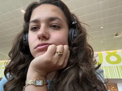 A close-up, selfie-style shot shows a young woman with long, dark, wavy hair, wearing black over-ear headphones. She is leaning on her hand, with her chin resting on her knuckles. Her expression is neutral, and her gaze is directed slightly off-camera. She is wearing a gold watch on her left wrist and a gold ring on her right middle finger. The background appears to be an indoor public space with a dropped ceiling and fluorescent lighting. There is a colorful graphic design on the wall behind her, featuring yellow and white stripes and a large yellow circular symbol. The environment suggests a modern, possibly commercial setting.