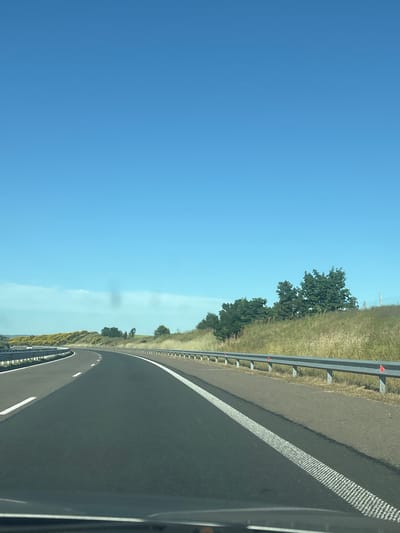 The image shows a clear blue sky over a highway in Vratitsa, Bulgaria. The highway curves to the left, with a metal guardrail on the right side and a dotted white line marking the lane. The surrounding landscape consists of grassy hills dotted with trees and yellow flowering bushes. The road surface appears to be asphalt. There are no people or vehicles visible in the immediate foreground.