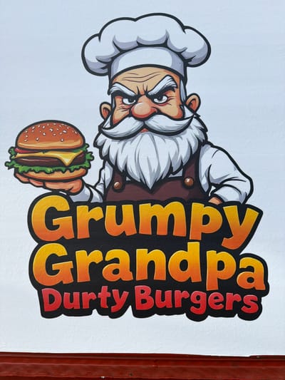 The image shows a brightly colored sign for "Grumpy Grandpa Durty Burgers". The sign features a cartoon illustration of a grumpy-looking old man with a white beard and chef's hat, holding a hamburger. The text is in large, bold letters with an orange and yellow gradient. The sign is mounted on a white wall, and a reddish-brown textured surface is visible at the bottom of the frame. The overall style is cartoonish and attention-grabbing. There are no specific details in the image that indicate a location in Gloucester, United Kingdom.