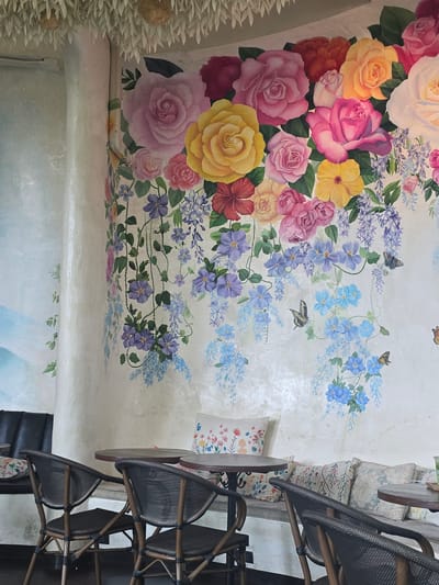 The image shows a restaurant ceiling with a floral mural in Ban Pa Daet North, Thailand. The mural features an array of colorful flowers, including roses and various blue and purple blooms, intertwined with butterflies. The restaurant's seating area is visible on the right side of the image, with chairs stacked against the wall. The overall aesthetic creates a garden-like atmosphere indoors.