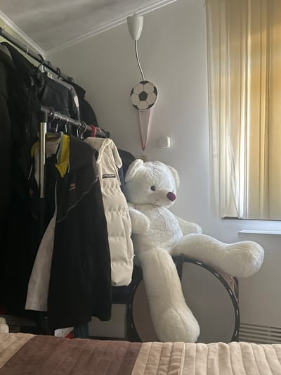 This image shows a cluttered indoor scene, likely a closet or storage area, in Madara, Bulgaria. The primary subjects are a large white teddy bear that is partly obscuring a dark, round object that appears to be a drum. The teddy bear is lying on its back, with its limbs splayed out. There are several pieces of clothing on hangers, including a white puffer jacket and black jackets, neatly organized on a metal rack. In the background, there are horizontal blinds with a soft, diffused light coming through them, suggesting it is daytime. A small white square object is mounted on the wall. To the left, a silver gooseneck lamp is attached to the wall, and a small black and white soccer ball is mounted on the wall above the lamp. The overall impression is one of a lived-in space, perhaps a child's room or a shared storage area. There is no activity taking place; the image captures a static scene. The visible text on the white jacket reads "HOODED COUTURE".