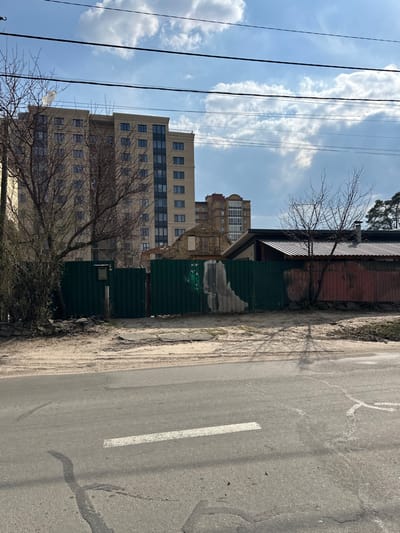 The image shows a street in Horenka, Ukraine (latitude: 50.525, longitude: 30.241) with a paved asphalt road in the foreground. The road is flanked by a sandy shoulder on each side. A green metal fence runs along the shoulder on the right side of the image. Behind the fence is a partially destroyed building with a red metal roof, a beige building, and a green tree on the left. The sky is partially cloudy. There is a white painted lane marker on the road. There are no people visible in the image. The scene appears peaceful and abandoned.
