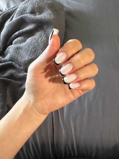 A close-up overhead shot shows a person's left hand with its fingers curled against a dark grey, wrinkled fabric. The hand has several nails that are painted with a French manicure style, featuring a sheer pale pink base and a sharp black tip. The fabric appears to be a blanket or throw. There is no identifiable location in the city of Rezh, Russia.
