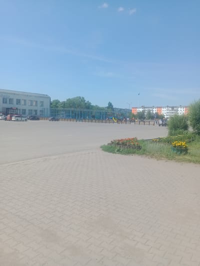 A wide-angle, eye-level outdoor shot depicts a paved area in Atbasar, Kazakhstan, under a clear blue sky with a few wispy clouds. The foreground features interlocking light-colored pavers. In the middle ground, a large, empty paved lot is visible, transitioning to a playground area with blue fencing and equipment, surrounded by trees. Beyond the playground, a low-rise building with a light-colored facade and windows is seen, with a sign that reads "Sinsay". Further in the background, there are apartment buildings, one with orange accents, and a tall flagpole flying a blue flag. Several parked cars are scattered across the paved lot. On the right side of the image, there are flower beds with red and yellow flowers and dense green bushes. A few individuals are visible near the playground and walking in the background.