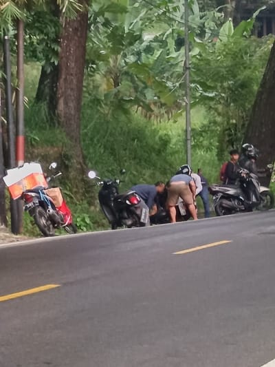 The image depicts a roadside scene in Cijedil, Indonesia. In the foreground, an asphalt road features a solid yellow line on the left and a segmented yellow line to the right. To the left of the road, a red scooter with a large white container and a brown cardboard box strapped to its rear is parked on an incline next to two dark vertical poles. Further right, a group of approximately five to six individuals are gathered. Three individuals are actively bent over, appearing to inspect or work on a black scooter parked by the road. One of these individuals wears a blue shirt, another tan shorts, and a third a white shirt. To their right, two other individuals stand near another black scooter; one wears a dark jacket and helmet, the other a dark top and red backpack. A fourth black scooter is also visible. The background consists of dense, lush green vegetation, including large-leaf plants and trees, suggesting a tropical environment.