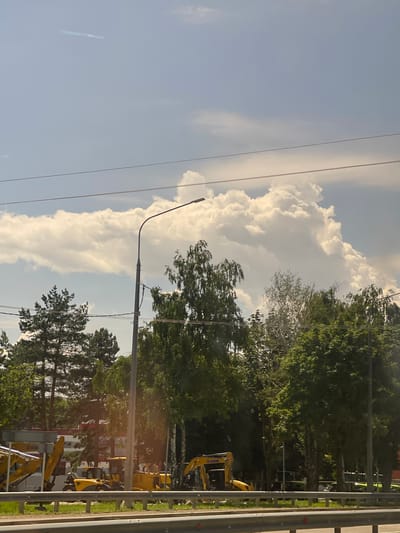The image shows a daytime scene in Верхнерусское, Russia, under a partly cloudy sky. The sky is a bright blue with large, fluffy white clouds dominating the left side of the frame. The sun is shining, casting a warm light on the scene.
In the foreground on the right, a metal guardrail with horizontal yellow and gray bars suggests a roadside or construction area. Beyond the guardrail, a line of lush green trees, including deciduous and coniferous varieties, forms a dense backdrop. The trees are vibrant and full, indicating a warm season.
Several yellow construction vehicles, likely excavators or backhoes, are visible among the trees and in the middle ground. Some are parked, while others appear to be in operation, suggesting ongoing construction or land development. A tall lamppost with a curved arm stretches across the sky from the lower left towards the center, its shadow faintly visible. Power lines crisscross the sky, adding to the urban or semi-urban feel of the location. In the background, a building with a light-colored facade and red accents can be seen peeking through the trees, hinting at a residential or commercial area.
The overall impression is one of a bright, active day in a developing or undergoing maintenance area, with nature and construction coexisting. There is no visible text in the image.