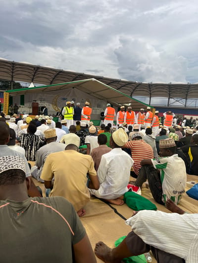 The image captures a wide-angle view of a large outdoor gathering of predominantly men at Amaan Stadium in Zanzibar City, Tanzania. The attendees are seated cross-legged on light brown woven mats, facing an elevated stage in the background. Many individuals wear traditional kufi caps, varying in color and pattern (white, beige, brown, black).

The stage is covered by a large white tent structure with green trim, featuring the "PBZ BANK" logo, a crescent moon and star motif, and the text "The People's Bank, For the People." On the stage, a dark wooden podium with a microphone is visible. Several men are present on or immediately in front of the stage; some wear white thobes and kufis, while others wear high-visibility safety vests (orange or yellow) over their clothing. One individual in a suit is visible behind the podium.

Beyond the stage structure, the curved, open-air upper tiers of stadium seating, colored blue, are discernible. The sky above is heavily overcast with dark grey clouds. The scene portrays a formal, large-scale event or ceremony.