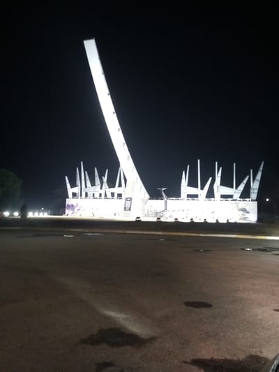 The image depicts the Campo Carabobo monument in Venezuela at night. The monument is illuminated and stands prominently against the dark sky. The foreground consists of a paved area with some minor debris or puddles. To the left, there is a glimpse of greenery and lights. A silhouetted figure can also be seen in the vicinity of the monument.