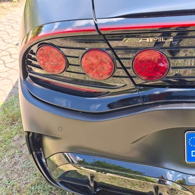 The image shows a close-up of the rear of a black car, likely a Mercedes-AMG, parked outdoors on a sunny day. The car features a prominent grill with vertical slats and three circular red taillights. A golden "AMG" emblem is visible on the grill. A European license plate with the EU stars and a blue background is also present. The car's paint reflects the surrounding environment, including trees and a cloudy sky. The ground is covered with grass and a paved area, suggesting a parking lot or driveway. No people are visible in the image, and there is no specific activity taking place beyond the car being parked. The lighting and reflections indicate it is daytime.