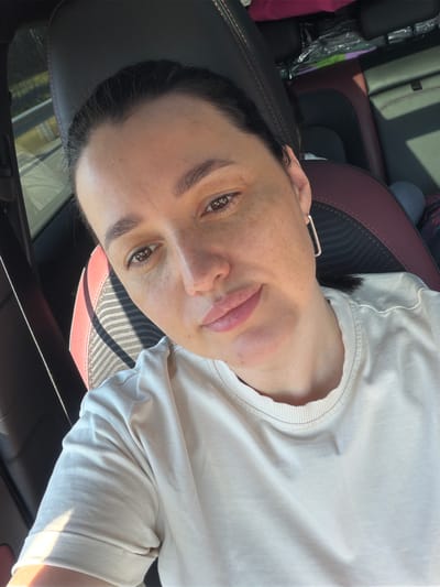 A woman with brown hair and brown eyes, wearing a light-colored t-shirt and square earrings, is taking a selfie in a car. The car's interior, featuring dark leather seats with red stitching, is visible. Sunlight casts shadows across the scene. The woman has freckles on her face. The location is identified as Uvarovo, Russia.