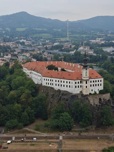 The image shows a panoramic view of the city of Děčín, Czechia, dominated by the Děčín Castle perched on a rocky outcrop. The castle, a large white building with a red tiled roof, features a prominent clock tower. Below the castle, a dense green forest covers the rocky terrain. In the background, the city of Děčín unfolds with various buildings, including residential structures and industrial facilities, one of which is marked by a tall, red-and-white striped smokestack. Highways with overpasses and a river are also visible, with a hazy mountain range forming the distant backdrop. The foreground reveals a park area with pathways, scattered people, bicycles, and some temporary event structures like tents and portable toilets, indicating a public gathering or event. The sky is overcast, contributing to a muted lighting condition.