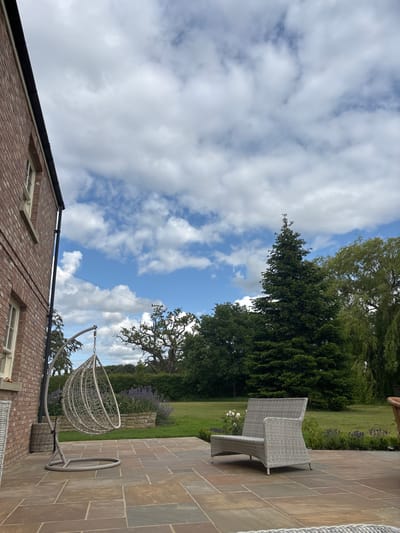 The image shows a patio in Sand Hutton, United Kingdom, located at coordinates (latitude: 54.025, longitude: -0.933). The patio is made of large, flat, brown stone tiles. There is a white wicker bench in the center of the patio, facing away from the viewer. A large hanging chair, constructed of woven gray material, sits on the left side of the image. A brick house is to the left of the image with a small window visible, and a stone pot with a leafy plant is on the lower left edge of the photo. In the background, a tall, dark green evergreen tree stands, surrounded by a row of other trees and foliage. Behind them is a grassy area and a hedge. The sky is blue with many white cumulus clouds.