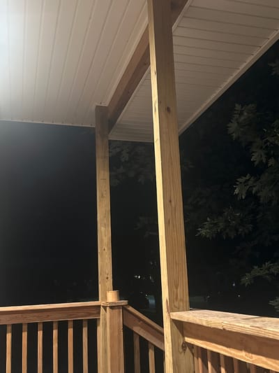 The image shows the underside of a wooden porch roof at night. The porch is supported by two vertical wooden posts and a horizontal wooden beam. The ceiling of the porch is white and paneled. In the background, there are dark trees with leaves and a dark sky. The porch railing is made of wood with vertical slats. The location is identified as Sedalia, United States of America. There are no people or animals visible in the image.