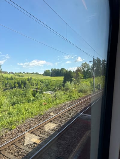The image shows a view from the window of a train traveling on railroad tracks in the Austrian town of Berg, Austria. The coordinates 47.84 latitude and 13.08 longitude confirm the location. The tracks are in the foreground, followed by a green, grassy embankment, and a grove of trees. In the distance, a small village with a few houses is visible behind the trees. The sky is blue with a few white clouds. There are electrical wires running parallel to the tracks.