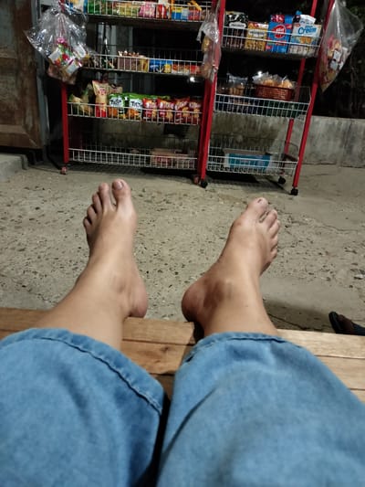 The image shows a person's bare feet and legs, clad in blue denim jeans, resting on a wooden surface. In the background, a red metal display rack is filled with various packaged snacks and goods. The scene appears to be outdoors, with a concrete ground and a wall visible. A portion of a dark-colored shoe or slipper is visible at the bottom right of the frame. The location is identified as Rantopanjang, Indonesia.