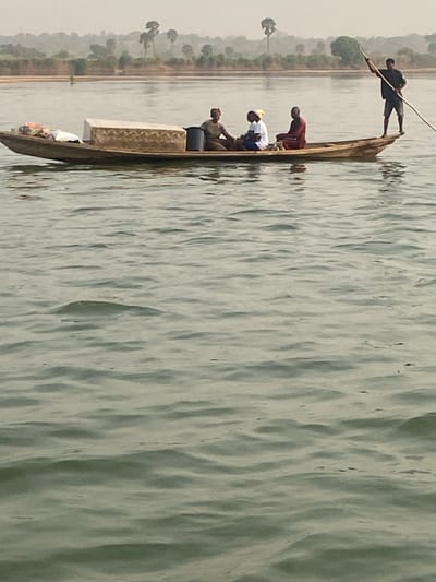 A narrow boat filled with people navigates a body of water, possibly a river or lake. One person stands at the bow, using a long pole to propel the boat forward. Three other individuals are seated inside. The water has gentle ripples and a light green hue. The shoreline, visible in the distance, is lined with sparse vegetation, including several tall, thin trees. The overall scene has a muted, overcast quality.