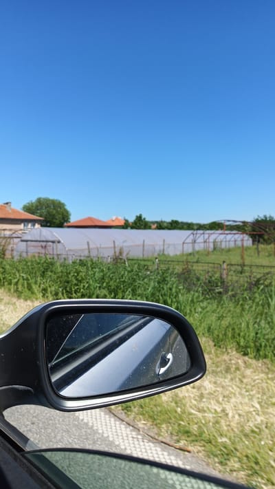 View from a car window of greenhouses and homes in Seltsi, Bulgaria. The rearview mirror of the car is visible in the foreground, reflecting the car door handle. Beyond the mirror is a field of tall green grass. In the background, multiple greenhouses stretch across the landscape. Further back, houses with orange tile roofs are visible, set against a clear blue sky.