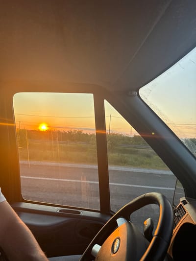 The image presents a perspective from inside a vehicle during what appears to be either sunrise or sunset. The primary subject is the sun, a brilliant yellow-orange orb, positioned low on the horizon and creating a significant lens flare that bathes the scene in warm light. The sky exhibits vibrant orange and yellow tones.

Through the car's windows, a road with white lane markings is visible, stretching into the distance. Beyond the road, a flat landscape with a line of distant trees or vegetation is silhouetted against the bright sky. The car's glass surfaces show some dust or marks.

Inside the vehicle, a black steering wheel, clearly displaying a "Ford" logo, occupies the right side of the frame. Elements of the dashboard, including an air vent, are also discernible. Part of a person's forearm, wearing a white short-sleeved shirt, is visible in the lower right, suggesting a driver or passenger is present. The car's interior roof lining and structural components frame the external view.

The scene conveys a sense of calm during the golden hour, likely as the vehicle is traveling or stopped on a road in the vicinity of Νέα Μεσολακκιά, Greece.