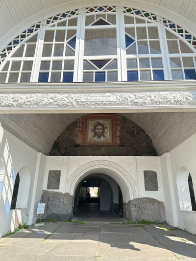 A medium shot captures the exterior of a white stone arched entryway in Solovetskiy, Russia, under a bright, sunny sky. The entryway is framed by large white stone arches. Above the entryway, a large, arched window with white mullions is visible, containing panes of clear, blue, and colored glass. A decorative white stucco frieze runs horizontally above the entryway, featuring grape and leaf motifs and Cyrillic text.

Centered above the inner archway, embedded in a brick niche, is a mosaic icon of Jesus Christ, rendered in the Byzantine style. Large, rough-hewn gray stones are placed on either side of the archway, supporting the structure. The floor is paved with large, irregular gray flagstones, with patches of green grass growing between them. Sunlight casts strong shadows across the pavement and the right side of the entryway.

Inside the archway, a dimly lit passageway recedes into the distance, with a single human silhouette visible in the far background, appearing to be walking away from the viewer. The walls of the passageway are visible, and a small white sign with text and graphics is mounted on a metal stand to the left of the entrance, in front of the large stone on the left. The overall impression is one of historical significance and religious importance, set against a backdrop of natural elements and architectural grandeur.
