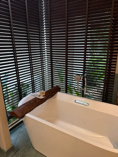 A bathtub with a wooden accent and a shower head is shown from a high angle. The bathtub appears to be made of a creamy white material and has a sleek, modern design. A dark brown, textured piece of wood is placed across the edge of the bathtub, and on it sits a small gold-colored object, possibly a ring. The shower head is made of polished metal and is connected to a faucet.
The scene is set in a bathroom with a unique ceiling structure. The ceiling is composed of parallel, dark brown slats, creating a slatted effect. Between these slats, glimpses of lush green foliage can be seen, suggesting the bathroom is located in a tropical environment, possibly with an outdoor or garden view. The lighting suggests it is daytime, with natural light filtering through the slats. The overall impression is one of a luxurious and serene spa-like setting, blending indoor comfort with the natural beauty of the outdoors. The location is identified as Ao Cho, Thailand.