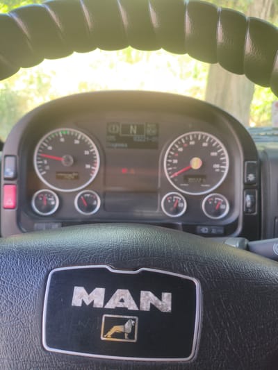 A close-up interior shot shows the dashboard and steering wheel of a MAN truck. The steering wheel, with a textured black leather grip, is prominently featured in the foreground, displaying the silver "MAN" logo with a lion emblem below it. Behind the steering wheel, the truck's dashboard is visible, featuring a circular speedometer with a red needle pointing to approximately 50, and a digital display showing "100022". Two smaller circular gauges are below the speedometer. To the right of the speedometer is a digital screen displaying what appears to be diagnostic information, including "086", "N", and "03221-05". Various buttons and controls are also visible on the dashboard. The background is blurred, showing greenery and sunlight filtering through trees, suggesting an outdoor setting. The image is taken from the perspective of someone seated in the driver's seat. The specific location in Alchevsk, Ukraine is not discernible from the image itself.