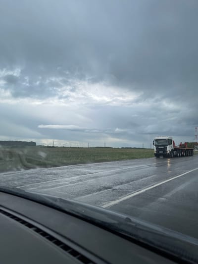 The image depicts a view from inside a vehicle, likely a car, looking out onto a highway. It appears to be a cloudy, possibly rainy day, indicated by the wet road surface and overcast sky. A large white truck, possibly a semi-trailer, is visible in the adjacent lane. There are grassy fields bordering the highway. Power lines can be seen in the distance. The location is Кокино, Russia.