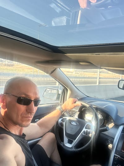 A bald, fair-skinned man wearing sunglasses and a black tank top is driving a car. He has a watch on his left wrist and a necklace around his neck. The car has a sunroof open, allowing sunlight to stream in. The interior of the car appears to be a modern SUV, with a Ford steering wheel visible. Outside the car, a highway can be seen, with a barrier and some vegetation. The sky is clear and blue. The lighting suggests it is daytime. The location is the al-Qasoum Regional Council in Israel, a predominantly desert area.