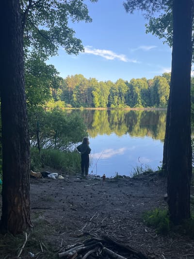 This image captures a serene outdoor scene, likely a lake or wide river, set in Krasnyi Jar, Russia. The photograph is taken from a vantage point framed by two large tree trunks, one at the bottom foreground and another at the top, creating a natural aperture through which the landscape is viewed.

The central feature is a calm body of water, reflecting the clear blue sky and a few wispy white clouds, as well as the dense green forest lining the distant bank. The reflections on the water are almost as vivid as the actual scenery, indicating tranquil conditions.

On the near bank, to the right of the frame, a person is visible, clad in dark clothing and with light-colored hair. They appear to be engaged in fishing, with at least one fishing rod extending into the water. Near the person, on the muddy and somewhat debris-strewn ground, are several items including a bottle and some white plastic bags or containers. The ground on this side is a mix of dark soil, sparse grass, and scattered twigs and branches.

The overall impression is one of peaceful recreation in nature. The bright blue sky with scattered clouds suggests a clear, pleasant day, likely morning or early afternoon, with ample sunlight illuminating the scene. There is no visible text in the image.