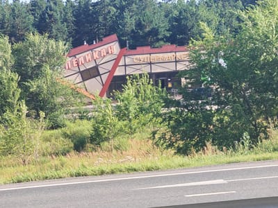 A tilted building with red roofing and a sign that reads "ПЕЛИКАН" in red letters is visible behind trees and foliage. To the right of this building, another section of the structure has a sign that reads "СУПЕРМАРКЕТ" in yellow letters. The building is set against a backdrop of dense green trees, possibly a forest. In the foreground, a paved road with white lane markings is visible, suggesting a highway or main road. The surrounding environment appears to be a rural or semi-rural area, with grassy slopes and bushes. The weather appears to be clear and sunny.