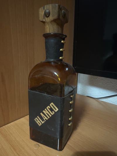 A unique brown glass bottle with a wooden stopper is displayed on a wooden surface. The bottle has a dark leather wrap around its base, featuring the word "BLANCO" in gold lettering at an angle. Gold decorative studs are visible along the sides of the leather wrap. The bottle stands in front of a wooden wall and a dark monitor screen. There is no indication of the location being Santa Pola, Spain.