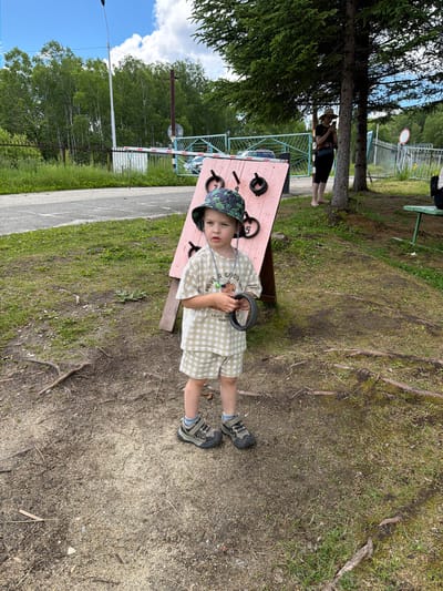 A young child stands on a dirt path in what appears to be an outdoor recreational area in Kul'dur, Russia. The child is wearing a checkered outfit, a patterned bucket hat, and sneakers. They are holding a black ring in their right hand. Behind the child is a pink wooden structure with various circular openings and some black rings attached. In the background, trees line a fence, and a gate with a barrier is visible. A woman is standing under a tree to the right of the frame, looking at her phone. The ground is covered in dirt, small stones, and patches of grass. The sky is blue with scattered white clouds.