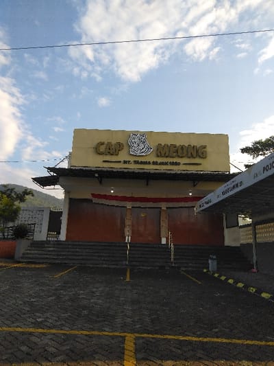 A frontal, static daytime image captures the "CAP MEONG" building and its immediate surroundings in Cijedil, Indonesia. The two-story building features a large yellow facade on the upper level displaying "CAP MEONG" in gold lettering with a white tiger head emblem, and "NY. TJONG OE JIEK 1888" below it. The ground level facade is brown, with a prominent set of wide, dark grey steps leading to closed brown entrance doors. A red and white banner, indicative of the Indonesian flag, hangs horizontally above the entrance. To the right, a dark, covered canopy extends over part of the parking area, while a white, patterned wall is visible to the left. The foreground consists of an empty parking lot paved with grey interlocking bricks, delineated by yellow painted lines. No individuals or vehicles are present. The background features a clear blue sky with scattered white clouds, and distant green mountains are visible on the left horizon.