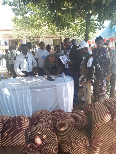 The image shows a group of people gathered around a white table draped in a white tablecloth, with a white sheet covering the table on one side. The table is set with a black box, a microphone, and two bottles of water. A man in a black suit and tie is standing at the table, signing something on a piece of paper. There are other people standing around the table, including a woman in a white shirt with a blue logo, a man wearing a white hat, and several other men and women, some wearing backpacks. The scene is outdoors in Bunia, Democratic Republic of the Congo, at the coordinates 1.556 latitude and 30.244 longitude. There is a light brown building behind them. Some of the people are standing under a tree with green leaves. In the foreground, there are three brown and cream striped cushions. The people are casually dressed in clothing ranging from formal to casual attire. The image appears to be a candid photo taken in a public space. The setting suggests that the group of people is engaged in a meeting or gathering. The image shows a group of people gathered around a table, with a man signing something on a piece of paper. The people are casually dressed and the scene appears to be a candid photo taken in a public space.